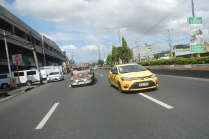 Ein gelbes Taxi fährt eine Straße mit hohen Gebäuden, Bäumen, Pflanzen, Polen, Lampen, Schildern und Kabeln entlang, unter einem bewölkten Himmel.