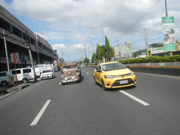Ein gelbes Taxi fährt eine Straße mit hohen Gebäuden, Bäumen, Pflanzen, Polen, Lampen, Schildern und Kabeln entlang, unter einem bewölkten Himmel.