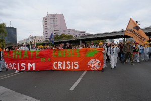 Eine Gruppe von Menschen marschiert öffentlich mit einem Banner, auf dem "Klima-Krise ist eine Krise" steht, mit Bäumen, Gebäuden und einer Brücke im Hintergrund bei einem bewölktem Himmel.