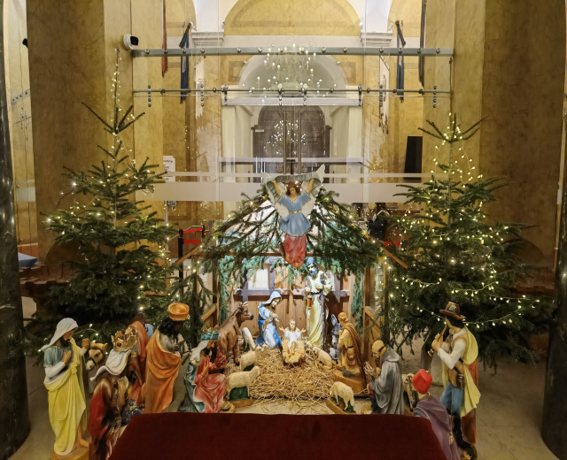 Krippe in einer Kirche mit einem roten Tuch auf einem zentralen Tisch, flankiert von zwei beleuchteten Weihnachtsbäumen und einer Kronleuchter über einem Geländer im Hintergrund.
