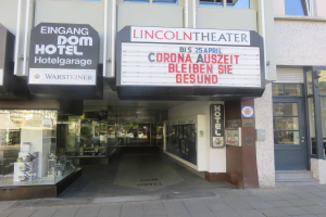 Das Lincoln Theater in Berlin, Deutschland - ein Gebäude mit Glasfenstern und -türen sowie einer Tafel mit Text, die verschiedene Objekte im Inneren zeigt und so den Eindruck einer pulsierenden Stadtkulisse vermittelt.