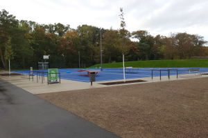 Ein Park mit einem Basketballplatz in der Mitte, umgeben von Straßenlaternen, Bänken und einem Mülleimer, mit grünem Gras, Bäumen und einem klaren blauen Himmel im Hintergrund.