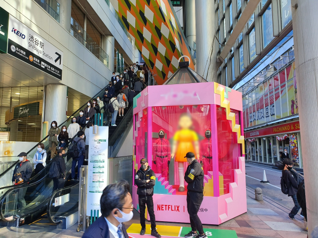 Eine Gruppe von Menschen steht vor einem Gebäude mit Glaswänden, Säulen und Texttafeln, einige tragen Masken, während eine Rolltreppe mit Fahrgästen zu sehen ist. In der Mitte steht ein pinker Kasten mit Schaufensterpuppen und der Aufschrift "Netflix's new Netflix booth in Tokyo."