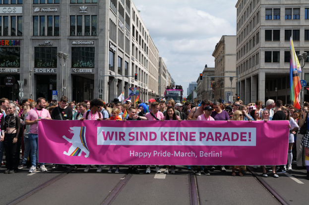 Eine Gruppe von Menschen marschiert auf einer Berliner Straße mit einem pinken 'Happy Pride March'-Schild, mit Gebäuden, Laternenmästen und Verkehrszeichen an der Straße.