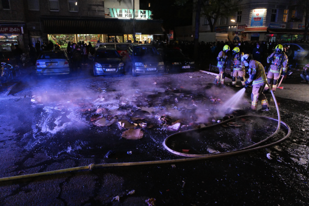 Eine Gruppe von Feuerwehrleuten in Helmen, die Rohre halten, steht um ein Straßenfeuer mit Fahrzeugen in der Nähe, Gebäuden, Schildern, Laternen, Bäumen und einem dunklen Himmel im Hintergrund.