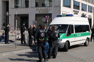 Eine Gruppe von Polizisten steht vor einem Starbucks-Café mit einem Van auf der rechten Seite und einigen Menschen in der Nähe, während im Hintergrund ein Gebäude mit Fenstern, einer Tafel, einem Laternenpfahl und einem Zaun zu sehen ist.