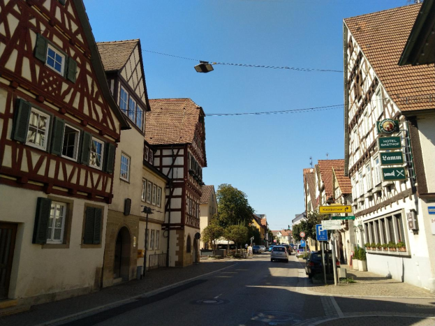 Eine Straße in Rothenburg ob der Tauber, Deutschland, mit fahrenden Autos, Gebäuden mit Fenstern und Schildern auf beiden Seiten und Bäumen mit einem klaren blauen Himmel im Hintergrund.