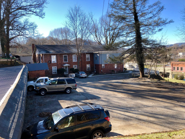 Ein Parkplatz mit geparkten Autos vor einem Backsteingebäude an der 609 W Main Street in Mount Vernon, VA, umgeben von Bäumen, Pflanzen und Gras, mit Hügeln und einem klaren blauen Himmel im Hintergrund und einer Wand auf der linken Seite.