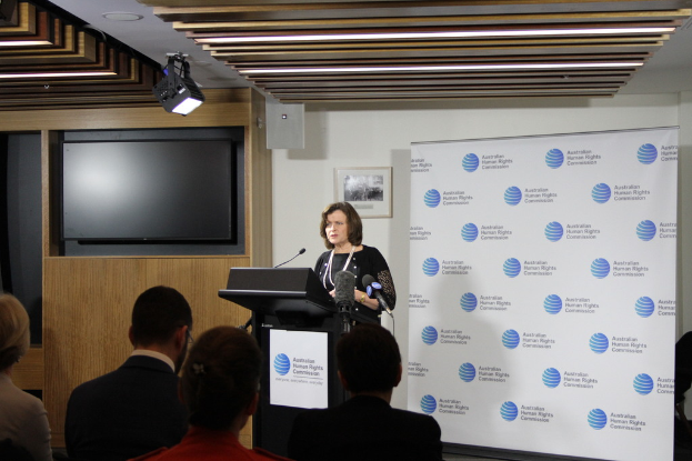 Eine Frau steht auf einer Bühne und spricht in ein Mikrofon, vor ihr sitzen Zuschauer, hinter ihr eine Wand mit einem Banner und ein Fernseher auf einem Holzwand, sowie Lampen an der Decke.