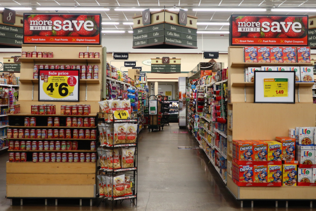 Ein Spätkauf-Interieur mit verschiedenen Lebensmitteln auf Regalen und dem Boden, Informationsschildern, Deckenleuchten und einer Person im Hintergrund.