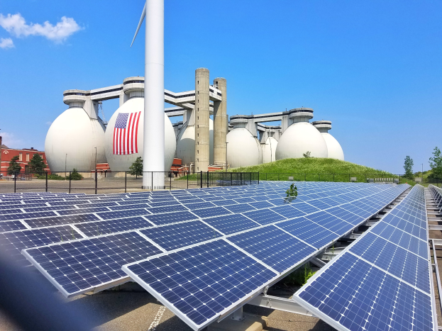 Solarpanels auf dem Dach eines Geb├Ąudes mit einer Flagge, umgeben von einem Zaun, Gras, Pflanzen, B├Ąumen, Polen und anderen Geb├Ąuden, mit einem bew├Âlktem Himmel im Hintergrund.