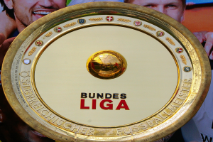 Goldener Schild mit "Bundesliga"-Text, gehalten von einem Mann mit einem Banner im Hintergrund, das drei Personen zeigt.