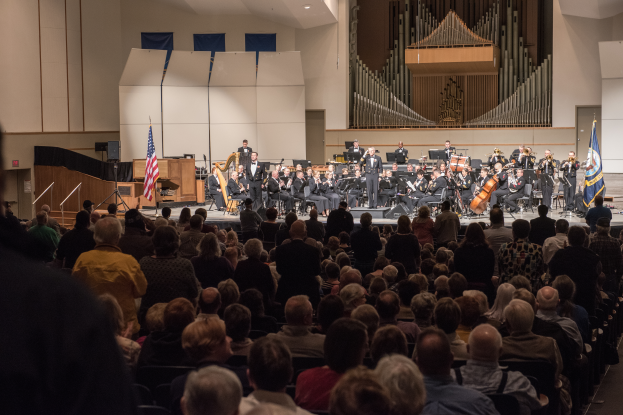 Eine große Gruppe von Menschen sitzt vor einer Bühne mit einigen, die Musikinstrumente halten. Flaggen mit Stangen, ein Lautsprecherbox und andere Gegenstände sind auf der Bühne zu sehen. An der Rückwand befindet sich eine Decke mit Lampen und eine Tür, was auf ein Konzert an der University of Texas at Austin hindeutet.