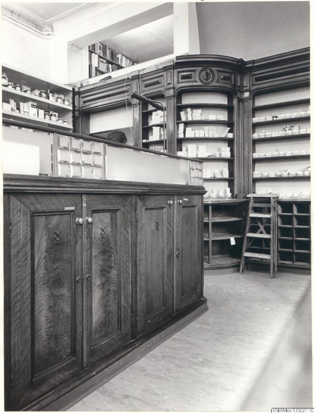 Schwarzes und weißes Foto eines vintage-apothekeninneren mit einer Theke, Schränken, einer Leiter und Regalen, die mit verschiedenen Gegenständen gefüllt sind, mit Text oben.