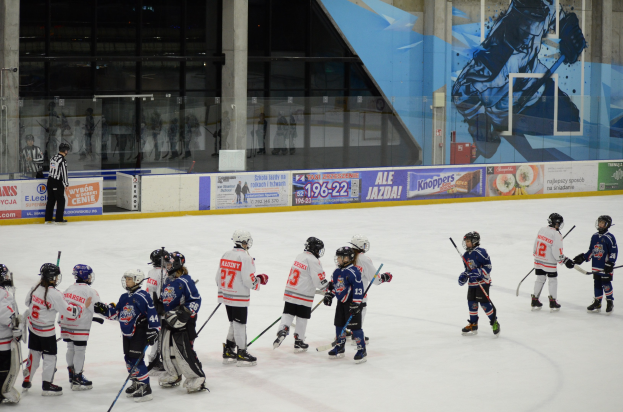 Junge Menschen in Helmen und Eishockey-Ausrüstung spielen Eishockey auf einer Indoor-Eisbahn mit Bannern, Säulen und Glaswänden im Hintergrund.
