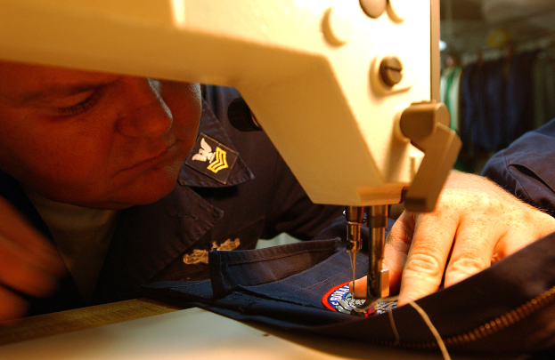 Ein Mann bedient eine Nähmaschine in einer Fabrik, konzentriert auf das Annähen von Stoff, mit Kleidung im Hintergrund.