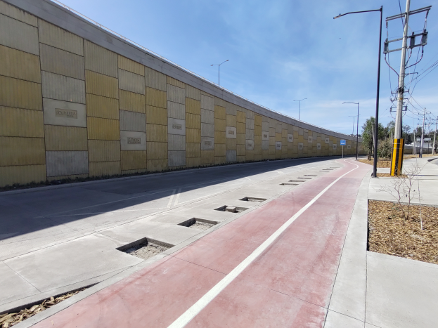 Ein Fahrradweg verläuft neben einer Straße, gesäumt von einer Mauer auf der linken Seite und Strommasten mit Drähten auf der rechten Seite, mit Bäumen im Hintergrund und einem bewölkten Himmel oben.