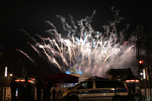 Ein Polizeiwagen steht vor einer Menschenmenge, im Hintergrund sind Gebäude, Bäume, Pfähle, Verkehrszeichen und Schilder beleuchtet, während farbige Feuerwerke den Himmel erhellen, während des Silvesterfeierns in Berlin.