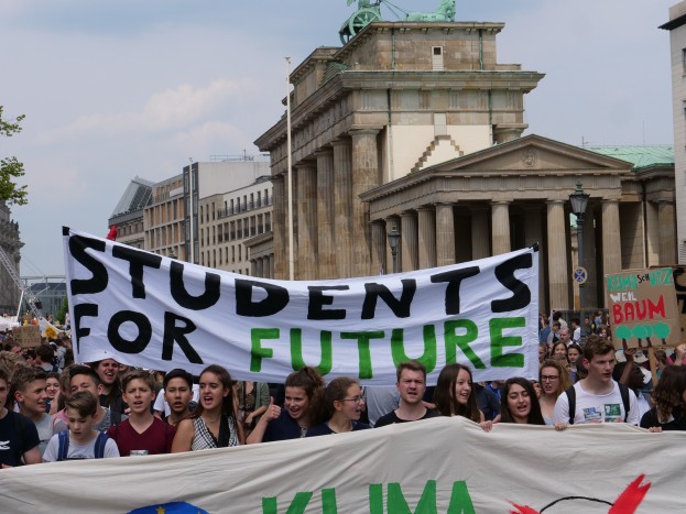 Eine Gruppe von Studenten marschiert in Berlin, die eine bunt bemalte Tafel mit der Aufschrift "Students for Future" trägt, mit Gebäuden, Bäumen und Himmel im Hintergrund.
