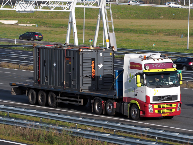 Ein Lastwagen mit einem großen Behälter fährt auf einer Autobahn, mit anderen Fahrzeugen, Pfählen, Bäumen, Gebäuden und einem klaren blauen Himmel im Hintergrund.