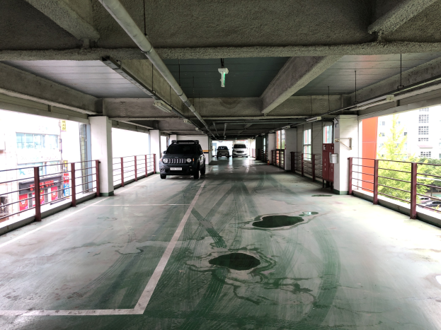 Parkhaus mit Autos zwischen Geländern, Säulen, Lampen und Rohren geparkt, umgeben von Bäumen und Stadtgebäuden mit Fenstern im Hintergrund.