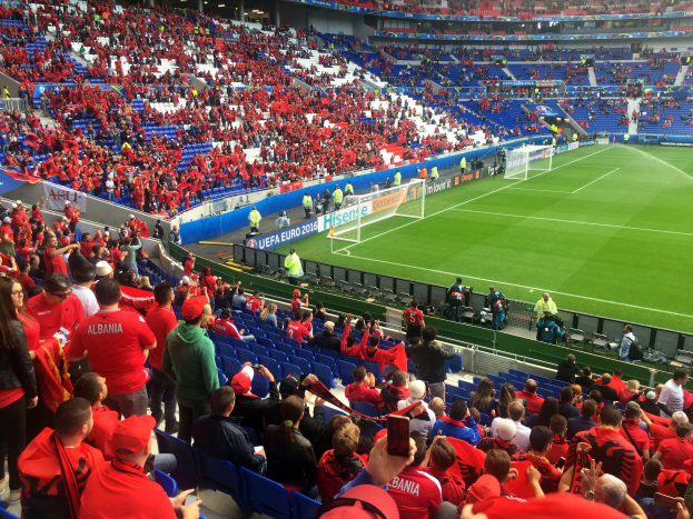 Ein volles Stadion mit Sitz- und Stehplätzen, Fahnen, ein Tor und Deckenbeleuchtung, während die Menge gespannt auf den Anpfiff des Fußballspiels wartet.