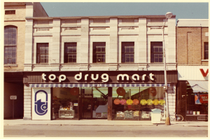 Ein Drogengeschäft an einer Straßenecke mit einem Gebäude, einem Schild, einem Straßenpfosten, einem geparkten Fahrrad, einem Müllcontainer und einem bewölkten Himmel.