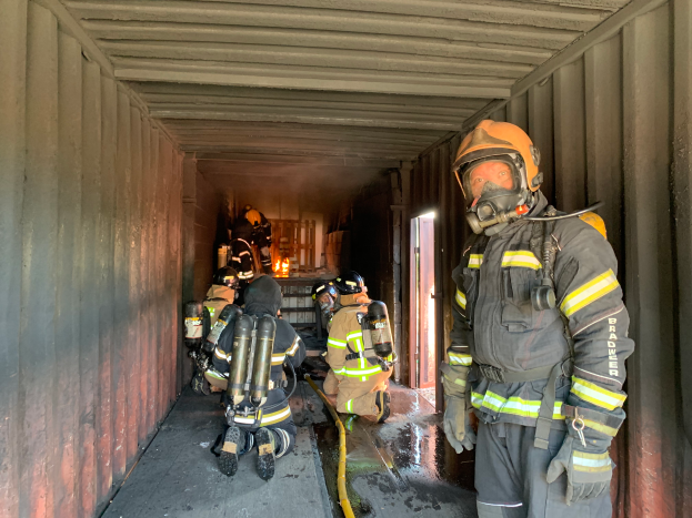 Eine Gruppe von Feuerwehrleuten in Helmen, Handschuhen und Sauerstoffzylindern steht in einem Seecontainer mit einem Rohr auf dem Boden, einer Tür im Hintergrund und anderen Gegenständen.