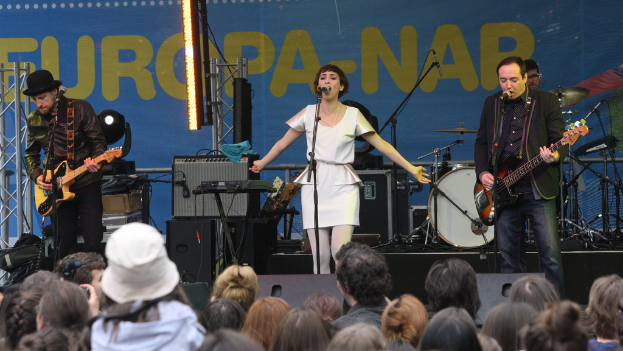 Eine Bühne mit vier Musikern: eine Frau in einem weißen Kleid singt in der Mitte, zwei Männer spielen Gitarren auf beiden Seiten und eine Menge ist unten zu sehen, mit einer Wand und Lautsprechern im Hintergrund.