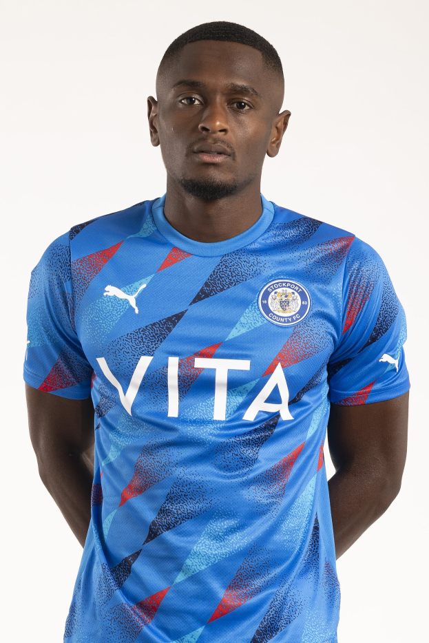Ein Mann in einem blauen Leicester City Away-Shirt der Saison 2018-19, das das Team-Logo, einen Rundhalsausschnitt und kurze Ärmel aufweist, steht mit den Händen in den Taschen und einem leichten Lächeln da.