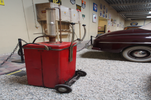 Eine rote Tankstelle auf Kies mit einem geparkten Auto rechts daneben, einer Wand mit Plakaten und einer Decke mit Lampen im Hintergrund.