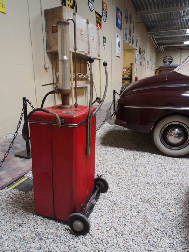 Eine rote Tankstelle auf Kies mit einem geparkten Auto rechts daneben, einer Wand mit Plakaten und einer Decke mit Lampen im Hintergrund.