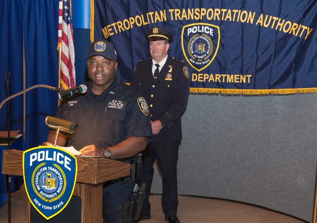 Zwei Polizeibeamte stehen vor einem Rednerpult, einer hält Papiere und spricht in ein Mikrofon, mit einem Banner, einer Flagge und einem Tisch in der Nähe, und einer Tafel mit Symbolen und Text im Hintergrund.