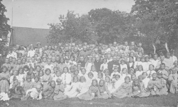 Schwarzes und weißes Foto einer Gruppe von Kindern, die vor einem Haus mit Bäumen und einem klaren Himmel posieren.