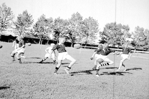 Schwarzes Foto von jungen Jungs, die auf einem Feld mit Bäumen und einem Pfosten im Hintergrund Fußball spielen.