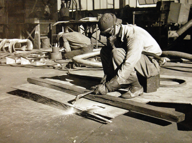 Ein Schweißer in einem Helm, Schutzbrille und Handschuhen arbeitet an einem Stück Holz in einer Fabrik, hält ein Schweißgerät und hat Rohre, Stangen und andere Gegenstände im Hintergrund.