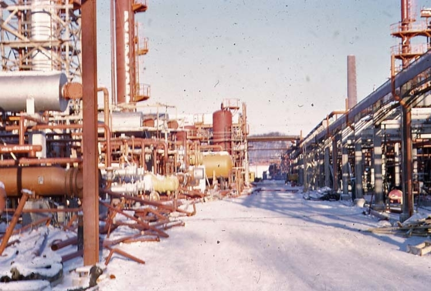Eine Ölraffinerie steht in der Mitte einer verschneiten Straße, umgeben von verschiedenen Maschinen und Mäständen, mit einer verschneiten Landschaft und einem Himmel im Hintergrund.