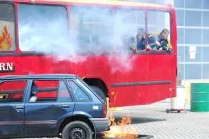 Roter Doppeldeckerbus mit Rauch und drei sichtbaren Passagieren, neben einem Auto geparkt, vor einem Gebäude mit Glasfenstern und einem Fass auf der rechten Seite.