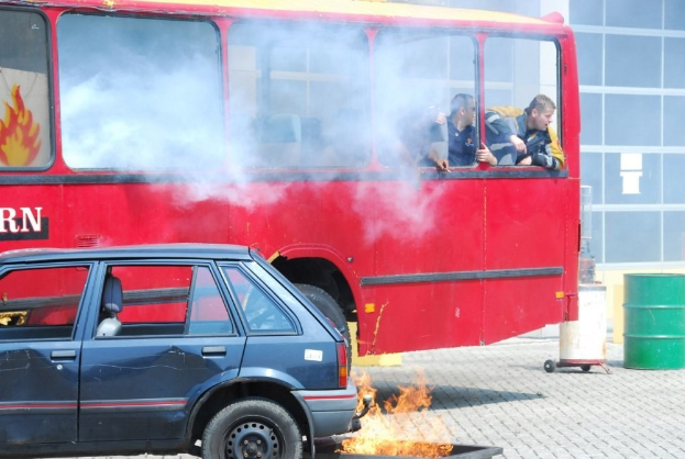Roter Doppeldeckerbus mit Rauch und drei sichtbaren Passagieren, neben einem Auto geparkt, vor einem Gebäude mit Glasfenstern und einem Fass auf der rechten Seite.