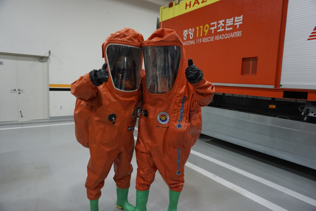 Zwei Personen in orangen Chemikalienschutzanzügen stehen nebeneinander vor einem LKW, mit einer Wand und Türen im Hintergrund, was darauf hinweist, dass sie Teil des Nationalen Einsatzteams sind.