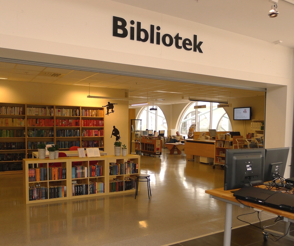 Mehrere hölzerne Regale voller Bücher, mit Monitoren auf der rechten Seite und dem Wort "BIBLIOTECA" oben angezeigt.