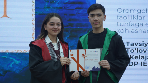 Ein Mann und eine Frau in Graduationsroben stehen Seite an Seite, jeder hält ein Zertifikat in der Hand, mit einem Bildschirm dahinter, der Text anzeigt.