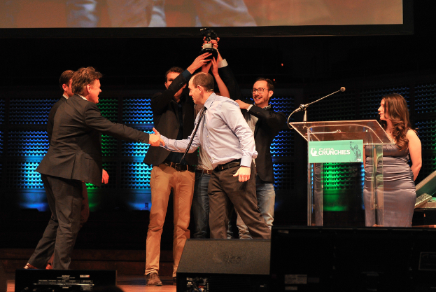 Zwei Männer in schwarzen Anzügen heben einen Preis mit den Händen und lächeln, während ein Mann links die Hand schüttelt und eine Frau rechts steht.