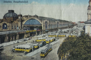 Eine alte Postkarte zeigt einen belebten Dresdner Bahnhof mit Fahrzeugen, Menschen, Bäumen, Gebäuden und einem bewölkten Himmel, mit der Aufschrift "Dresden Hauptbahnhof" oben.