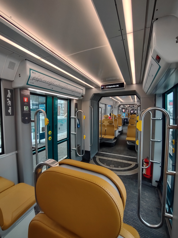 Das Innere eines Zuges mit gelben Sitzen, Metallstangen, Glastüren, Schilden, Deckenleuchten und einem sichtbaren Feuerlöscher.