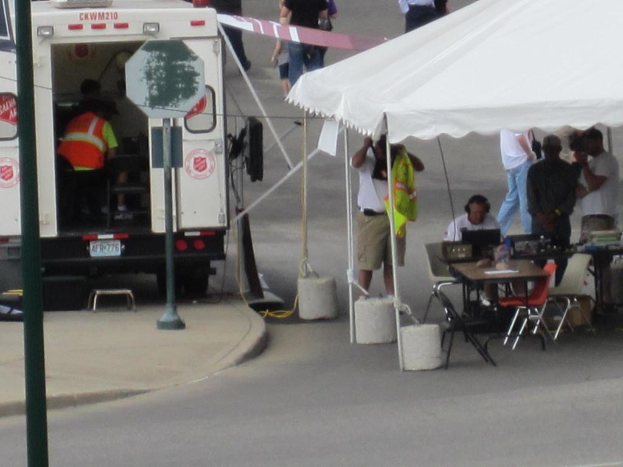 Ein Rettungswagen steht auf der linken Seite einer Straße, während auf der rechten Seite ein Zelt eine Gruppe von Menschen schützt, bei der ein Tisch und ein Stuhl in der Nähe stehen.