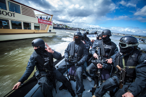 Eine Gruppe von Polizisten in schwarzen Uniformen und Helmen, einige bewaffnet, fahren auf einem Boot im Wasser mit einer Brücke, Bäumen, Gebäuden und einem klaren blauen Himmel im Hintergrund.