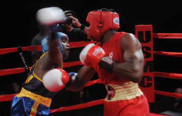 Zwei Boxer kämpfen im Ring mit einem Fotografen und Zuschauern im Hintergrund bei dramatischer Beleuchtung.