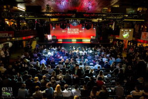 Eine große Menge sitzt auf Stühlen vor einer Bühne mit Bildschirmen und Bannern, die von Scheinwerfern beleuchtet und von Geländern umgeben ist, mit einem Wasserzeichen in der linken unteren Ecke, das eine Atmosphäre von Spannung und Erwartung schafft.
