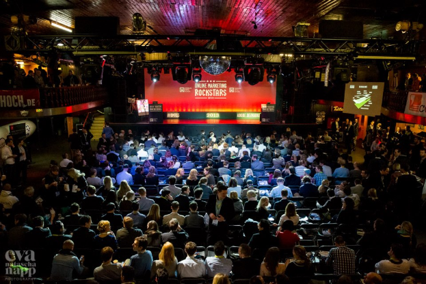 Eine große Menge sitzt auf Stühlen vor einer Bühne mit Bildschirmen und Bannern, die von Scheinwerfern beleuchtet und von Geländern umgeben ist, mit einem Wasserzeichen in der linken unteren Ecke, das eine Atmosphäre von Spannung und Erwartung schafft.